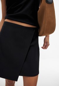 Gonna nera avvolgente realizzata in tessuto liscio, con un taglio pulito e una sovrapposizione diagonale. Una borsa in suede color cammello è portata di lato.