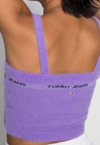 Fioletowy, puszysty crop top z cienkimi ramiączkami. Zawiera logo w postaci taśmy z napisem "TOMMY JEANS" w kolorze czarnym. Miękka faktura, szeroki elastyczny dół.