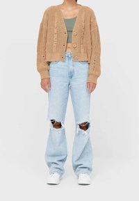 Beige gebreide cardigan met een losse pasvorm, voorzien van een gevlochten textuur en grote knopen, gecombineerd met oversized lichtblauwe versleten jeans.