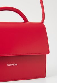 Raudona odinė rankinė, struktūrizuotos formos, su viršutine rankena ir sklende. Papuošta iškilniu "Calvin Klein" logotipu ir kontrastinga siūle.