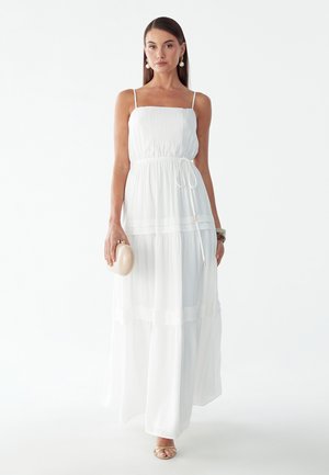 Vrouw draagt een witte mouwloze maxi-jurk, houdt een beige clutch vast, met hangende oorbellen en beige open-toe hakken, staand op een witte achtergrond.