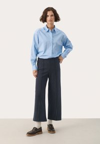Világoskék, hosszú ujjú ing, sötét csíkozott, bő szárú nadrággal párosítva. Barna fűzős cipő egészíti ki az outfitet.