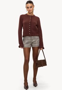 Pull en maille marron avec ourlet à volants et détail à boutons, associé à un short à carreaux. Le mannequin tient un petit sac à main marron et porte des escarpins pointus.