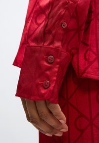 Manșeta unei cămăși roșii cu o textură subtilă, având un design cu model. Sunt vizibili trei nasturi roșii, iar materialul are un finisaj lucios.