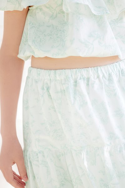 Conjunto de tecido com padrão floral branco, apresentando um top cropped com babados e uma saia de cintura elástica em verde claro, com uma textura suave e leve.