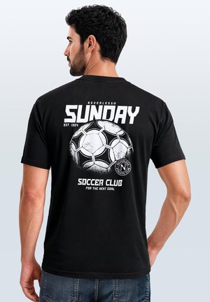 Mann, der wegschaut, trägt ein schwarzes T-Shirt mit weißem Fußball-Grafik und dem Text "Sunday Soccer Club" auf dem Rücken.