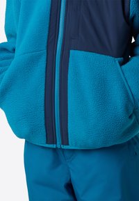 Τζάκετ από φλις σε τυρκουάζ με ναυτικό μπλε πάνελ, διαθέτει κεντρικό φερμουάρ και πλαϊνές τσέπες. Λείο υφή και αντίθετες λεπτομέρειες στις ραφές.