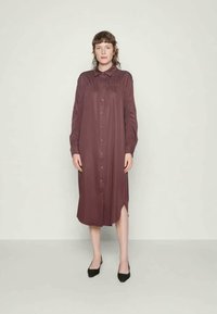 Mulher em pé contra uma parede simples, vestindo um vestido camiseiro de manga longa, botão, na cor bordô, e sapatos pretos de bico fino.