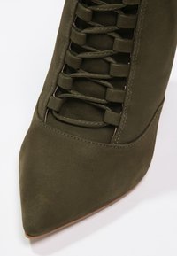 Botín de ante verde oliva de tacón alto con punta afilada, que presenta un diseño de cordones y una textura suave. El tacón es elegante y contorneado.