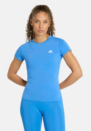 Frau mit lockigem Haar trägt ein eng anliegendes blaues Adidas-Shirt mit kurzen Ärmeln und passende blaue Sportleggings, steht mit den Händen hinter dem Rücken.