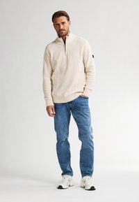 Pullover zip in maglia beige con un motivo testurizzato, indossato sopra jeans in denim scuro. Il modello si trova su uno sfondo bianco con calzature neutre.