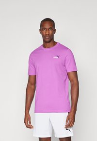 Lilla bomulds T-shirt med rund hals og korte ærmer; har et lille sort Puma-logo på venstre overdel. Båret med hvide shorts.