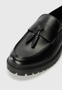 Black leather loafers with a smooth finish, featuring a broad band across the upper and two tassels. Thick, textured black rubber sole.