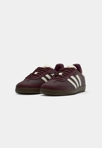 Et par mørk burgunder Adidas Samba sneakers med hvide striber, gummisåler og burgunder snørebånd på en hvid baggrund.