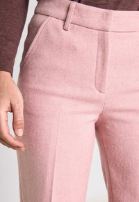 Pantalons ajustés rose clair en tissu texturé, dotés de poches latérales et d'une taille lisse. Main positionnée près de la cuisse.