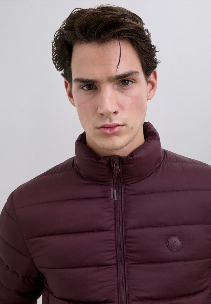 Chaqueta de plumón burdeos con patrón acolchado horizontal, cuello alto y cierre de cremallera. Presenta un logotipo redondo en el lado izquierdo del pecho.