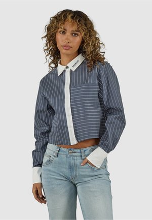 Mujer con cabello rizado que lleva una camisa corta de botones azul marino con rayas finas, puños y cuello blancos, combinada con jeans azul claro, de pie.