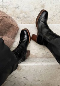 Stivaletti neri in pelle con texture a coccodrillo, punta arrotondata e tacco in legno. Posati su una superficie in marmo, con una morbida texture beige nei dintorni.