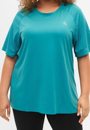 Femme portant un t-shirt de sport ample à manches courtes turquoise avec des manches en mesh et un pantalon noir, debout, les bras détendus le long du corps.
