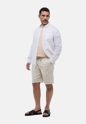 Chemise blanche en lin à boutons sur un t-shirt beige, associée à un short beige clair avec cordon de serrage et des sandales noires, se tenant devant un fond uni.