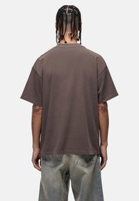 Bruine oversized t-shirt met korte mouwen en een ronde hals, van achteren weergegeven. De stof lijkt glad met een subtiele textuur.