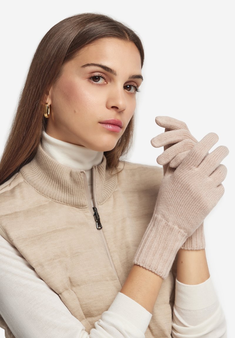 Guanti lavorati a maglia di colore rosa chiaro con polsini a coste. Il modello indossa un gilet beige con zip sopra un dolcevita bianco. Texture liscia, design aderente.
