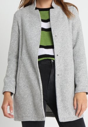 Lysegrå uldfrakke med trykknapper båret åben over en grøn, hvid og sort stribet sweater og sorte bukser med høj talje.
