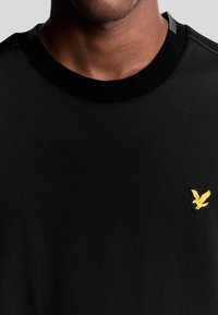 Felpa nera con scollo rotondo, con un logo giallo a forma di aquila sul lato sinistro e dettagli grigi sulle cuciture delle spalle.