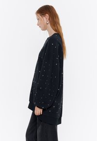 Sudadera negra de manga larga, corte oversized, con pequeños puntos multicolores y un dobladillo de canalé. Textura de tela suave.