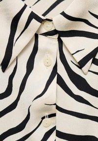 Gros plan sur un col de chemise crème avec des rayures zèbre noires et trois boutons crème alignés sur le devant.