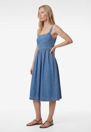 Kvinde iført ærmeløs blå denimkjole med firkantet halsudskæring og knælængde samt brune flade sandaler, stående med den ene hånd på hoften og kigger mod venstre.