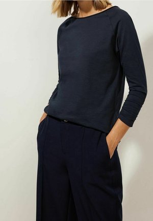 Vrouw draagt een marineblauwe top met lange mouwen en bijpassende wijde broekspijpen, handen in de zakken, tegen een effen lichte achtergrond.