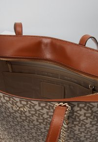 Sac fourre-tout marron avec une surface texturée, présentant un motif de logo. À l'intérieur, une poche zippée et un porte-cartes. Accents en métal chromé visibles.