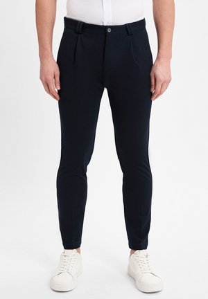 Navyblauwe slim-fit broek met plooien, een knoopsluiting en zijzakken, gecombineerd met witte sneakers op een lichte achtergrond.