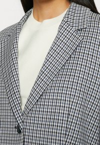 Blazer con un patrón de cuadros azul y blanco, solapas afiladas y un detalle de un solo botón, combinado con una blusa de punto de color claro debajo.