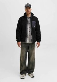 Zwarte fleecejas met een marineblauw zakje, grijs geruit overhemd en baggy vervaagde spijkerbroek. Schoeisel: lichtgrijze sneakers met donkere accenten.