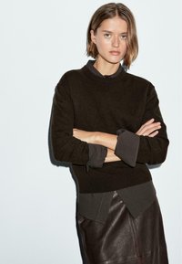 Maglione marrone scuro con scollatura rotonda, tessuto texture. Indossato sopra una camicia scura con colletto, stratificato con una gonna in pelle marrone.
