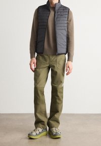 Grå puffer väst över en brun stickad tröja, kombinerad med olivgröna cargobyxor och grå sneakers med gröna sulor.