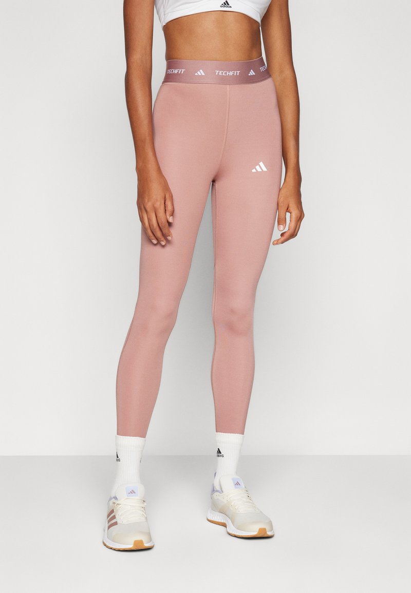 Leggings ajustées roses avec une taille élastique portant l'étiquette "TECHFIT" et des accents de logo blancs, associés à des chaussettes blanches jusqu'à la cheville et des chaussures de sport beiges.