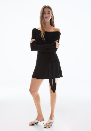 Mladá žena stojaca so založenými rukami, oblečená v čiernych dlhých šatách s odhalenými ramenami a bielych sandáloch na opätku na bielom pozadí.