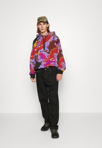 Hoodie met een meerkleurig barok patroon in paars, rood en goud; gecombineerd met zwarte broek en zwarte sportschoenen. Hoed met gouden accenten.