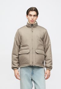 Beige polstret jakke med høj krave, frontlynlås og to store lommer. Elastiske manchetter og kant. Kombineret med lyseblå jeans.