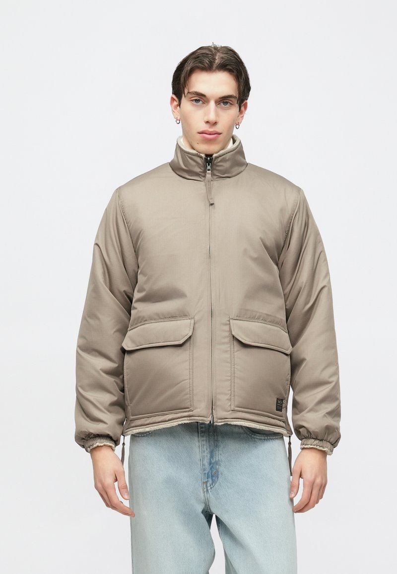 Beige polstret jakke med høj krave, frontlynlås og to store lommer. Elastiske manchetter og kant. Kombineret med lyseblå jeans.