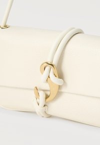 Τσάντα χειρός από κρεμ δέρμα με λουράκι που σχηματίζει θηλιά, στερεωμένο με λαμπερό χρυσό γάντζο, σε λευκό φόντο.