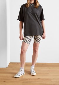 Persona con una camiseta negra holgada, pantalones cortos de ciclismo con estampado de cebra, zapatillas de deporte gris de caña alta y un collar de cadena plateada, de pie sobre un suelo de madera.