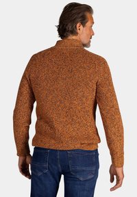 Gebreide trui in oranje en zwart gemêleerd garen, met een hoge hals en geribbelde manchetten. Draagt bij blauwe spijkerbroeken en zichtbare achterdetails.