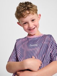 Jonge jongen met krullend haar glimlacht en wijst naar een gestreept roze en paars sportshirt met het "hummel" logo.