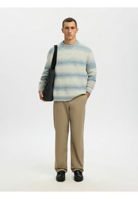 Jeune homme debout portant un pull rayé bleu clair, un pantalon beige, des chaussures noires et un sac à bandoulière noir.