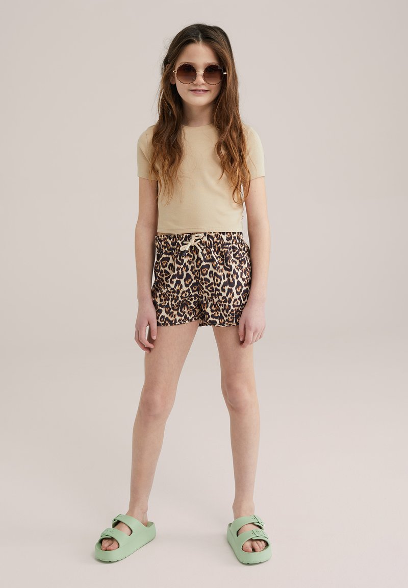 Beige T-Shirt mit kurzen Ärmeln, Shorts mit Leopardenmuster und Zugband, grüne Slipper mit verstellbaren Riemen, lange wellige Haare und Sonnenbrille.