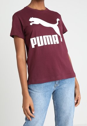 Maroon cotton t-shirt with a large white Puma logo. Short sleeves and a relaxed fit. Paired with blue denim jeans.